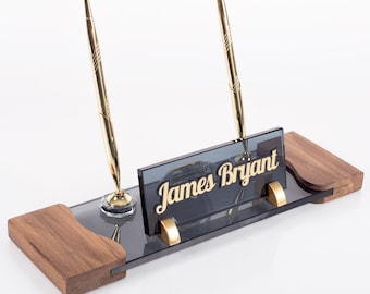 Könnte beinhalten: Ein Schreibtisch-Organizer aus Holz mit einem schwarzen Glas-Namensschild, auf dem "James Bryant" steht. Das Namensschild hat goldene Schrift. Zwei goldene Stifte befinden sich in den Haltern.