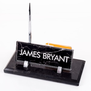 May include: Black marble desk nameplate with silver lettering that reads "JAMES BRYANT". A silver pen is in the holder on the nameplate.