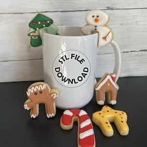 Puede incluir: Una taza de cerámica blanca con el texto "STL FILE DOWNLOAD" impreso en el lateral. La taza está decorada con varias galletas con temática navideña, incluyendo un muñeco de nieve, un árbol de Navidad, una casa de jengibre, un bastón de caramelo y un hombre de jengibre.