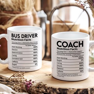 Peut inclure: Deux mugs en céramique blanche avec du texte noir. L'un dit "BUS DRIVER Nutrition Facts" et l'autre "COACH Nutrition Facts". Les mugs sont sur une surface en bois avec des éléments décoratifs en arrière-plan.
