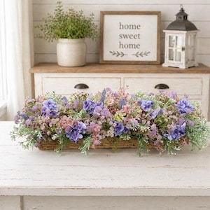 May include: A rectangular floral arrangement in a woven basket. The arrangement features a mix of purple, lavender, and pink flowers with green foliage. The basket sits on a white table, with a sign that says "home sweet home" in the background.