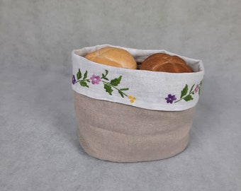 May include: A beige linen bread basket with a white embroidered trim featuring purple flowers and green leaves. The basket is filled with two rolls of bread.