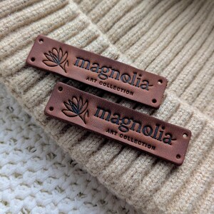 Puede incluir: Dos etiquetas de cuero marrón con la palabra "magnolia" y "ART COLLECTION" en relieve en negro. Cada etiqueta presenta un diseño floral. Las etiquetas están colocadas sobre un gorro de punto beige, con una superficie de punto blanco en el fondo.