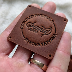 Puede incluir: Una etiqueta cuadrada de cuero marrón con el texto en relieve "RECYCLED MATERIALS" en el borde superior. El centro presenta la palabra "Samay" en una fuente estilizada, con "JEANS" debajo. El texto "RYKOVA TATYANA" está en relieve en el borde inferior.
