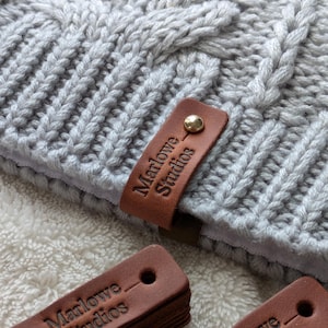 May include: Close-up of a light gray knitted hat with a cable knit design. A brown leather tag with the text "Marlowe Studios" is attached with a gold-colored rivet. Additional leather tags are visible.