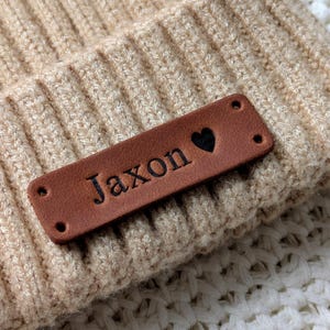 Peut inclure: Gros plan d'un bonnet en tricot beige avec une étiquette en cuir marron. L'étiquette porte le nom "Jaxon" et un symbole de cœur noir. Le bonnet a une texture côtelée.