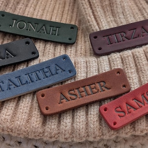 May include: A selection of personalised leather name tags in colours such as green, black, blue, brown, and red. Each tag has a different name embossed in capital letters. The tags are displayed on a beige knitted surface.