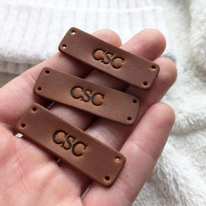 Puede incluir: Tres etiquetas de cuero marrón con las letras "CSC" grabadas. Cada etiqueta tiene dos agujeros en cada extremo. Se sostienen en una mano, con una tela blanca texturizada en el fondo.
