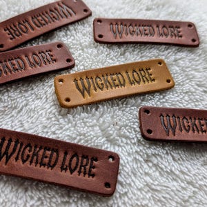 May include: Close-up of several rectangular leather tags with the words "WICKED LORE" embossed on them. The tags are in various shades of brown and tan, with two small holes on each end. The tags are scattered on a white, textured surface.
