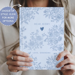 May include: A light blue journal with floral illustrations and the text "DEAR AVERY" and "LOVE, GRANDMA". The journal has a heart symbol and the text "CHOICE OF STYLE. CLICK FOR MORE OPTIONS" in a circle.