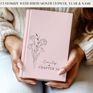 May include: A pink journal with a floral design and the text "Emily Chapter 30." The journal is personalized with the text "Customize with birth month flower, year & name" at the top. The journal is held in someone's hands.