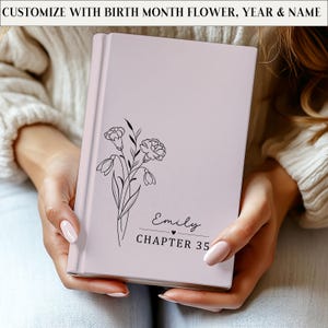 May include: A light pink journal with a floral design and the name "Emily" and "CHAPTER 35" printed on the cover. The journal is held in someone's hands. The text "CUSTOMIZE WITH BIRTH MONTH FLOWER, YEAR & NAME" is printed at the top.