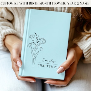 May include: A light blue journal with a floral design and the name "Emily" and "Chapter 21" printed on the cover. The text "Customize with birth month flower, year & name" is printed at the top. The journal is held in someone's hands.