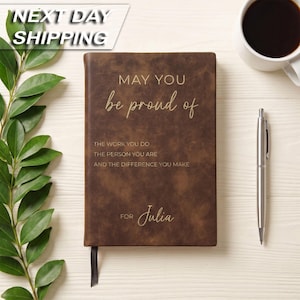 May include: A brown leather-like journal with gold text that reads: MAY YOU be proud of THE WORK YOU DO THE PERSON YOU ARE AND THE DIFFERENCE YOU MAKE FOR Julia. The journal includes a dark brown ribbon bookmark and is positioned next to a silver pen.