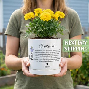 May include: A white ceramic planter containing bright yellow marigold flowers. The pot features the text Chapter 70, the name Fiona, a small purple flower graphic, a heart icon, the year EST. 1956, and a paragraph about memories and new dreams.