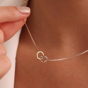 May include: A delicate necklace featuring two interlocking rings, one gold-toned and one silver-toned, suspended from a fine chain. The necklace is held up, showcasing the contrast between the metals. The jewelry is designed for everyday wear.