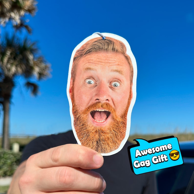 Pu&ograve; includere: Un profumatore per auto stampato con un uomo sorpreso con la barba rossa. Il profumatore ha un fumetto blu con il testo "Awesome Gag Gift" e un'emoji sorridente con gli occhiali da sole.