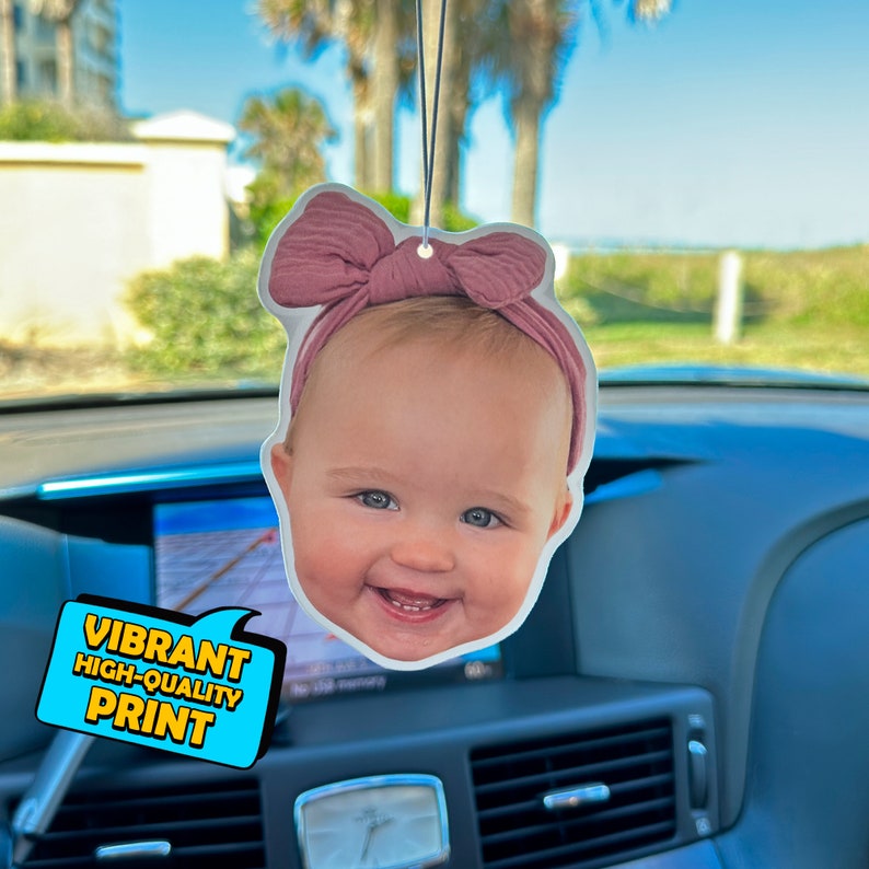 Pu&ograve; includere: Un profumatore per auto con una foto di un bambino con un fiocco rosa. Il profumatore &egrave; appeso allo specchietto retrovisore di un'auto. Il testo "VIBRANT HIGH-QUALITY PRINT" &egrave; visibile nell'angolo inferiore sinistro dell'immagine.