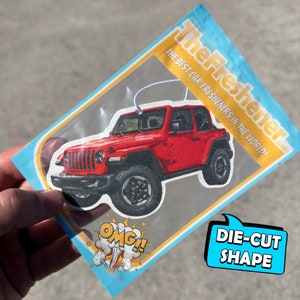 K&ouml;nnte beinhalten: Ein roter Jeep Wrangler Auto-Lufterfrischer in einer durchsichtigen Plastikt&uuml;te mit blauem und gelbem Rand. Die T&uuml;te tr&auml;gt den Text "The Freshener.com" und "The Best Car Fresheners in the World!" Der Lufterfrischer ist in Form eines Jeep Wrangler ausgeschnitten.