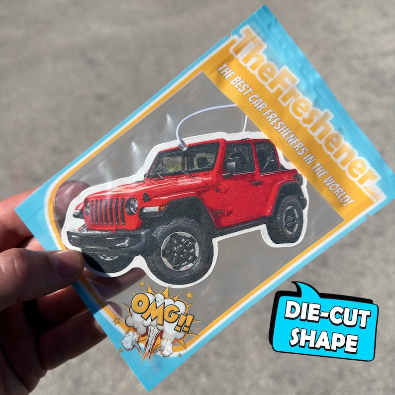 K&ouml;nnte beinhalten: Ein roter Jeep Wrangler Auto-Lufterfrischer in einer durchsichtigen Plastikt&uuml;te mit blauem und gelbem Rand. Die T&uuml;te tr&auml;gt den Text "The Freshener.com" und "The Best Car Fresheners in the World!" Der Lufterfrischer ist in Form eines Jeep Wrangler ausgeschnitten.