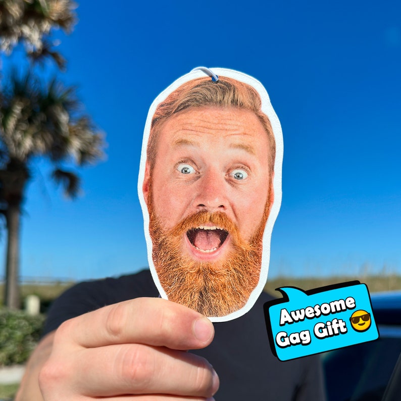 K&ouml;nnte beinhalten: Ein bedruckter Auto-Lufterfrischer mit einem &uuml;berraschten Mann mit rotem Bart. Der Lufterfrischer hat eine blaue Sprechblase mit dem Text "Awesome Gag Gift" und ein l&auml;chelndes Emoji mit Sonnenbrille.