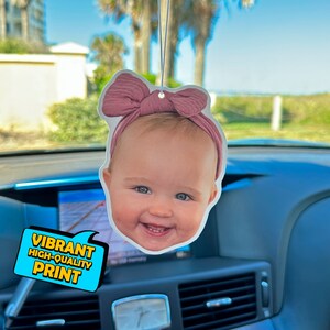 K&ouml;nnte beinhalten: Ein Auto-Lufterfrischer mit einem Foto eines Babys mit einer rosa Schleife. Der Lufterfrischer ist wei&szlig; mit einem lebendigen, hochwertigen Druck.
