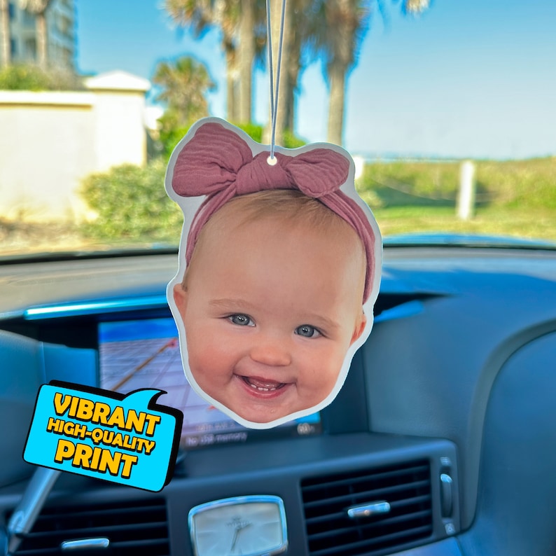 K&ouml;nnte beinhalten: Ein Auto-Lufterfrischer mit einem Foto eines Babys mit einer rosa Schleife. Der Lufterfrischer ist wei&szlig; mit einem lebendigen, hochwertigen Druck.