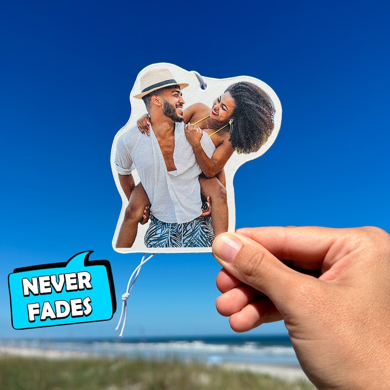 K&ouml;nnte beinhalten: Ein wei&szlig;er Auto-Lufterfrischer mit einem Foto eines l&auml;chelnden Paares. Der Mann tr&auml;gt einen Hut und die Frau einen Badeanzug. Der Text "NEVER FADES" befindet sich auf einer blauen Sprechblase.