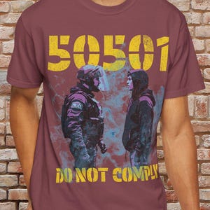 Peut inclure: Un t-shirt marron avec un imprimé graphique jaune de deux personnages, l'un avec un casque de riot et l'autre avec une veste à capuche. Le texte "50501 DO NOT COMPLY" est imprimé sur le t-shirt.