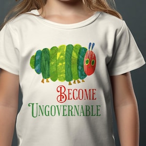Puede incluir: Una camiseta blanca con un diseño de oruga colorida y el texto "Become Ungovernable" en rojo y verde. La camiseta la lleva una niña pequeña con el pelo largo y rubio, sonriendo a la cámara.