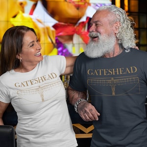 May include: Two people wearing t-shirts with the word "GATESHEAD" and a line drawing of a bridge. One shirt is white, the other is navy blue. The image shows the people smiling and embracing. The shirts appear to be made of cotton.
