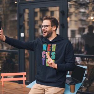 May include: A man wearing a black hoodie with the word "NEWCASTLE" spelled out in colourful letters. He is smiling and taking a selfie with his phone.