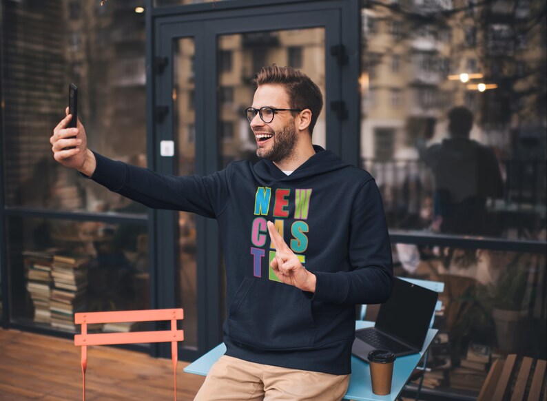 May include: A man wearing a black hoodie with the word "NEWCASTLE" spelled out in colourful letters. He is smiling and taking a selfie with his phone.