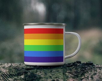 May include: A white enamel camping mug with a rainbow flag design. The mug is sitting on a tree trunk.