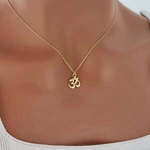May include: A gold-colored necklace featuring a small Om symbol pendant. The pendant is a stylized representation of the sacred Hindu symbol. The necklace is worn against a white top.