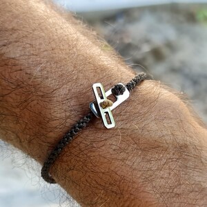 Peut inclure: Un bracelet tressé gris foncé avec un charm de couleur argentée. Le charm a une forme rectangulaire avec une petite pierre de couleur claire au centre. Le bracelet est porté au poignet.