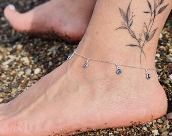 Könnte beinhalten: Silbernes Fußkettchen mit mehreren blauen bösen Augen-Anhängern. Das Fußkettchen befindet sich am Knöchel einer Person.