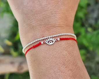 May include: Two bracelets are shown. One is a silver beaded bracelet. The other is a red string bracelet with a silver eye charm. The bracelets are worn on a wrist.