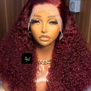 Op de afbeelding: Een bordeauxrode krullende pruik op een mannequinhoofd. De pruik heeft een kanten voorkant en een natuurlijke haarlijn. Een gouden kettingketting siert de nek van de mannequin. De pruik heeft een volumineuze, krullende textuur en is gestyled met een zijscheiding.