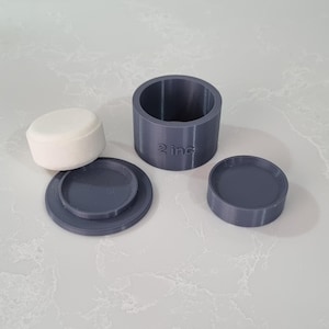 Może przedstawiać: Zdemontowany, ciemnoszary zestaw pojemników drukowanych w 3D. Zestaw zawiera cylindryczny pojemnik z napisem "2 inch", dwie okrągłe pokrywki i białą, cylindryczną gąbkę. Elementy są ułożone na jasnoszarej powierzchni.