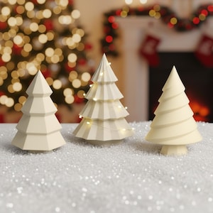 Puede incluir: Tres adornos de árbol de Navidad de color crema. Un árbol está iluminado con pequeñas luces de hadas. Los árboles están sobre una superficie blanca y brillante, con un árbol de Navidad y una chimenea borrosos en el fondo.