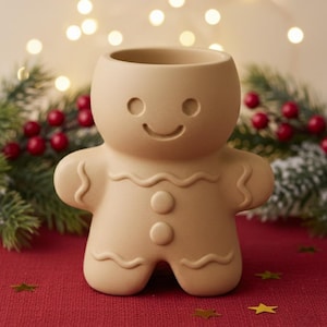 May include: A tan ceramic gingerbread man-shaped container with a smiling face, arms, and legs. The container has a large opening at the top. The background includes Christmas decorations and lights.