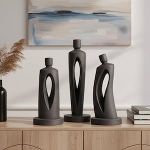 May include: Three dark gray abstract sculptures with a minimalist design. Each sculpture has a unique shape with a large, central opening. They are displayed on a light wood cabinet, with a painting in the background.