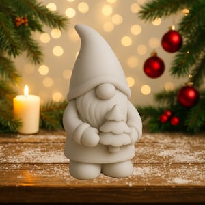 Op de afbeelding: Een wit kabouterbeeldje met een kleine kerstboom. De kabouter heeft een lange puntmuts, een ronde neus en een baard. Het beeldje staat op een houten oppervlak met een kaars en kerstversieringen op de achtergrond.