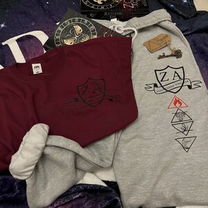 May include: A burgundy crew neck sweatshirt and grey sweatpants with matching graphic designs. The designs include the letters "ZA" within a shield, the words "AQUA TERRA IGNIS AER", and elemental symbols. A book and a key tag are also visible.