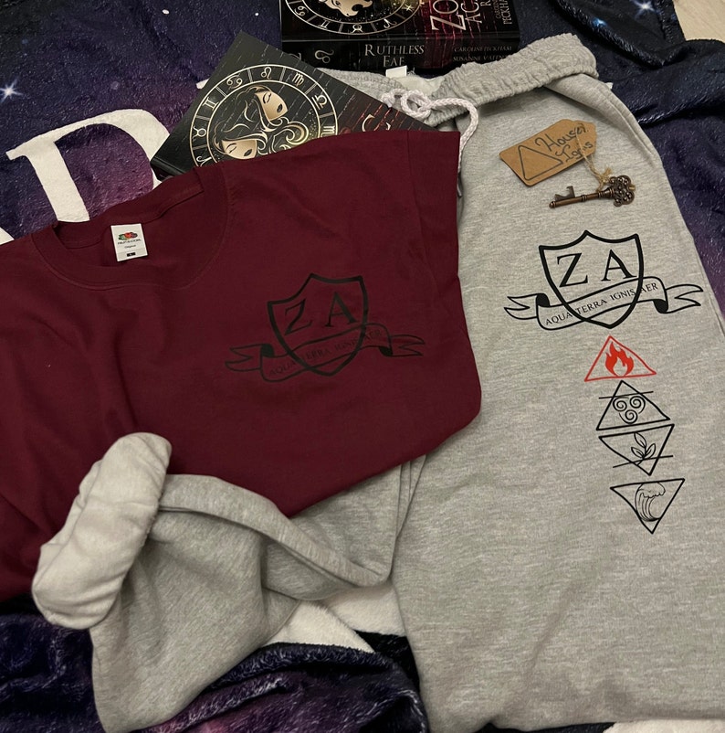 May include: A burgundy crew neck sweatshirt and grey sweatpants with matching graphic designs. The designs include the letters "ZA" within a shield, the words "AQUA TERRA IGNIS AER", and elemental symbols. A book and a key tag are also visible.