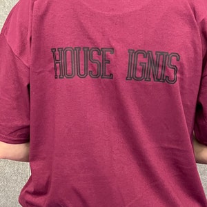 May include: A burgundy short-sleeved t-shirt featuring the text "HOUSE IGNIS" in black block letters on the back. The shirt is a solid colour and appears to be made of a soft material.