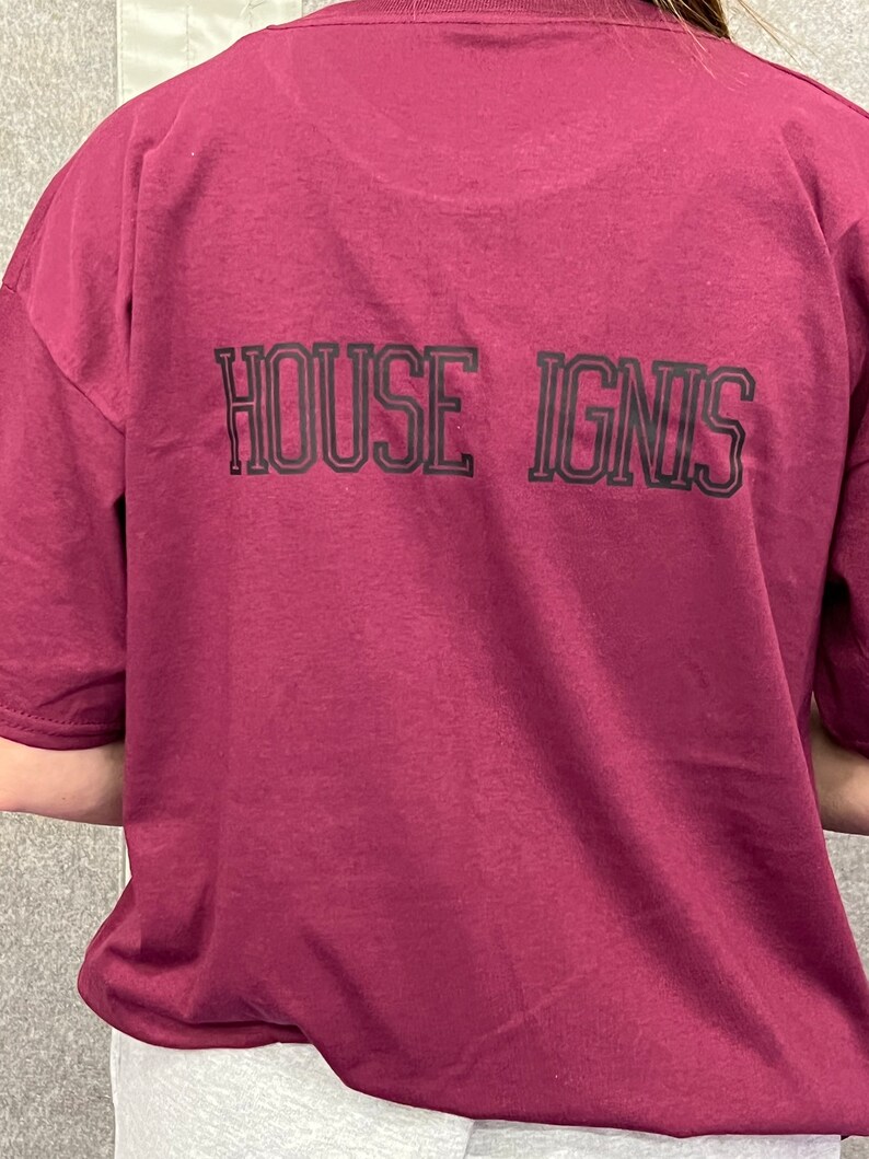 May include: A burgundy short-sleeved t-shirt featuring the text "HOUSE IGNIS" in black block letters on the back. The shirt is a solid colour and appears to be made of a soft material.