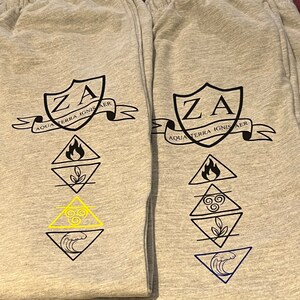 May include: Two pairs of grey sweatpants with elasticated waistbands. Each pair features a black crest with "ZA" and the text "AQUA TERRA IGNIS AER". Below the crest are four geometric symbols representing elements, with one pair having a yellow symbol.