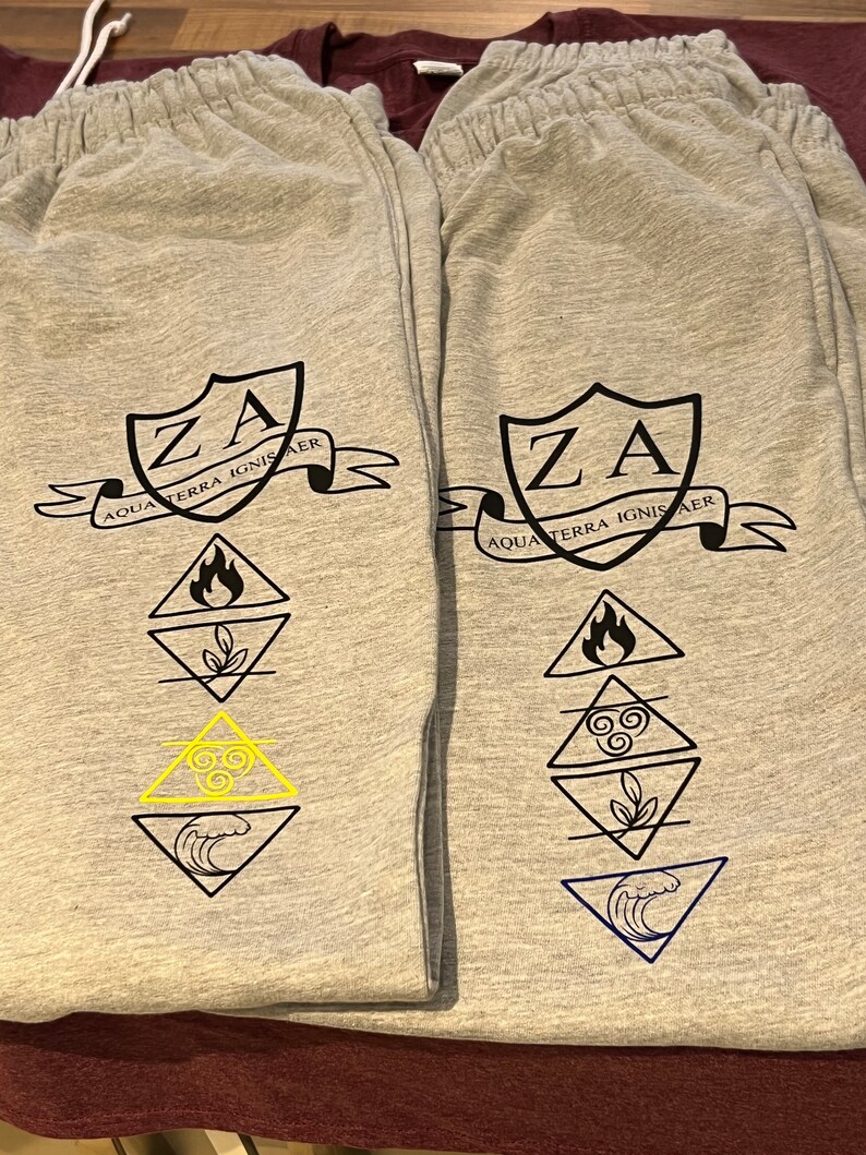 May include: Two pairs of grey sweatpants with elasticated waistbands. Each pair features a black crest with "ZA" and the text "AQUA TERRA IGNIS AER". Below the crest are four geometric symbols representing elements, with one pair having a yellow symbol.
